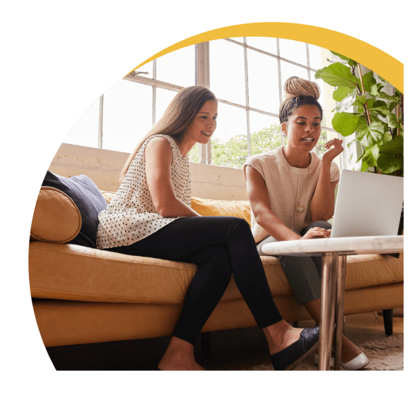 Two women working on a laptop while seating on sofa