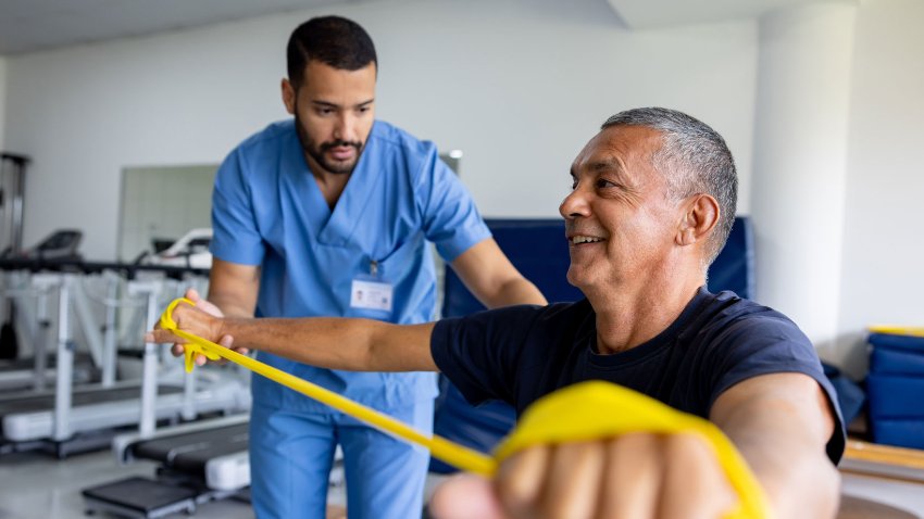 A Physiotherapist in blue scrubs helping man extend his arms using yellow stretching band