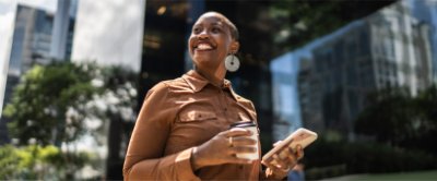 A woman standing outside in the city smiling while holding a coffee and a phone