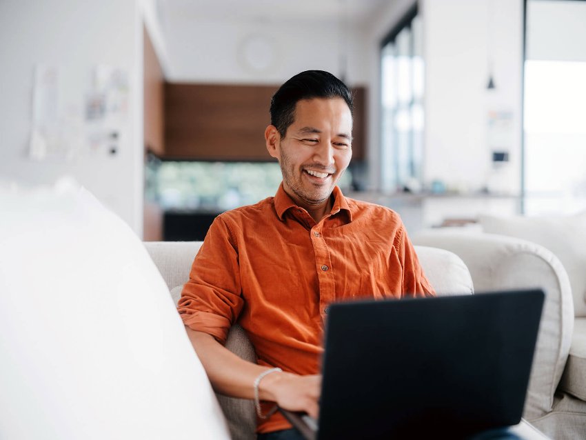 A happy man sitting on a couch, working or browsing the internet on his laptop from home